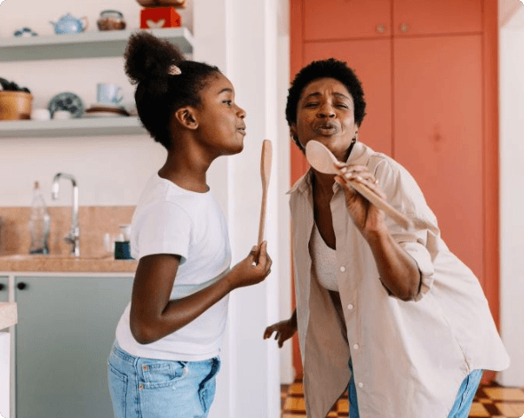 A happy grandmother and granddaughter playfully using spoons as microphones while singing and cooking in the kitchen. 