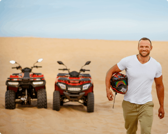 A person holding a helmet and smiling with two ATVs parked in the background.